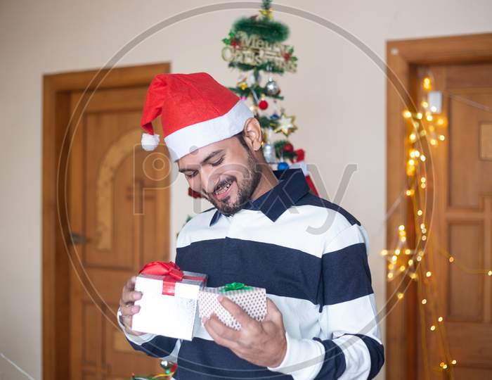 Happy Indian Man Wearing Santa Hat Holding Christmas Gift Boxes Or Presents Enjoys Holiday Season At Home,