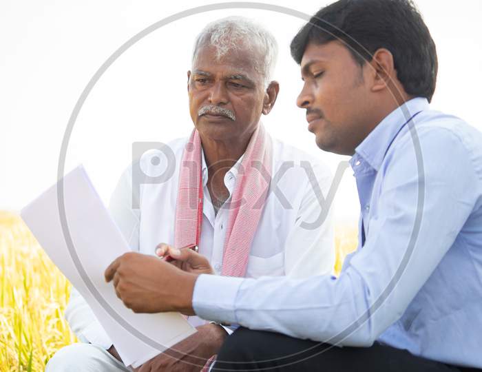 Selective Focus On Farmer, Bank Officer Explaning About Farm Loan Paper Documents To Indian Farmer Near Agriculture Field - Concpet Of Indian Villger Banking And Lifestyle.