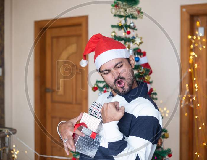 Cheerful Indian Man Wearing Santa Hat Holding Christmas Gift Boxes Or Presents Enjoys Holiday Season At Home, Extreme Happiness, Emotions, Copy Space