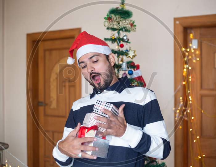 Cheerful Indian Man Wearing Santa Hat Holding Christmas Gift Boxes Or Presents Enjoys Holiday Season At Home, Emotions