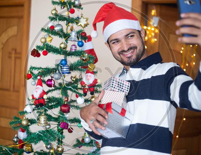 Happy Indian Man Wearing Santa Hat Talking Selfie Or Video Call Over Smart Phone Holding Christmas Gift Boxes Or Presents Enjoys Holiday Season At Home, Christmas Decoration Tree In Background.