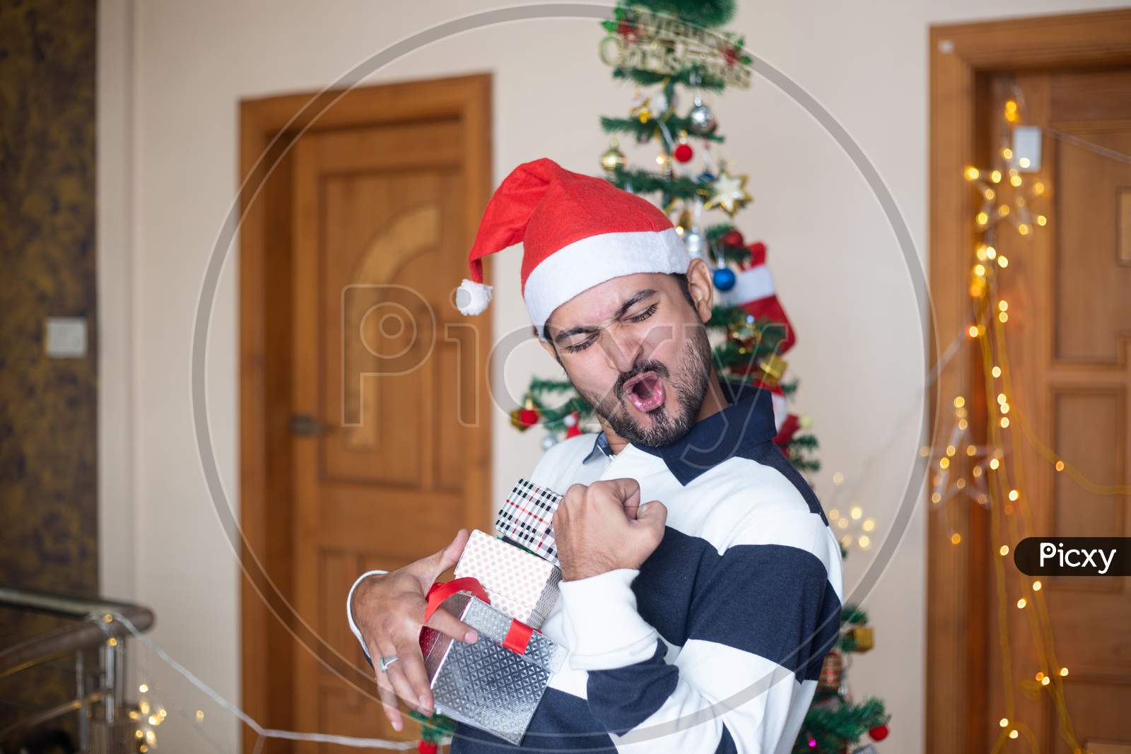 Cheerful Indian Man Wearing Santa Hat Holding Christmas Gift Boxes Or Presents Enjoys Holiday Season At Home, Extreme Happiness, Emotions, Copy Space