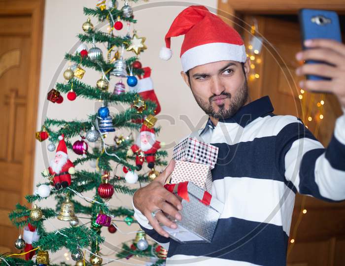 Happy Indian Man Wearing Santa Hat Talking Selfie With Smart Phone Holding Christmas Gift Boxes Or Presents Enjoys Holiday Season At Home, Christmas Decoration Tree In Background.
