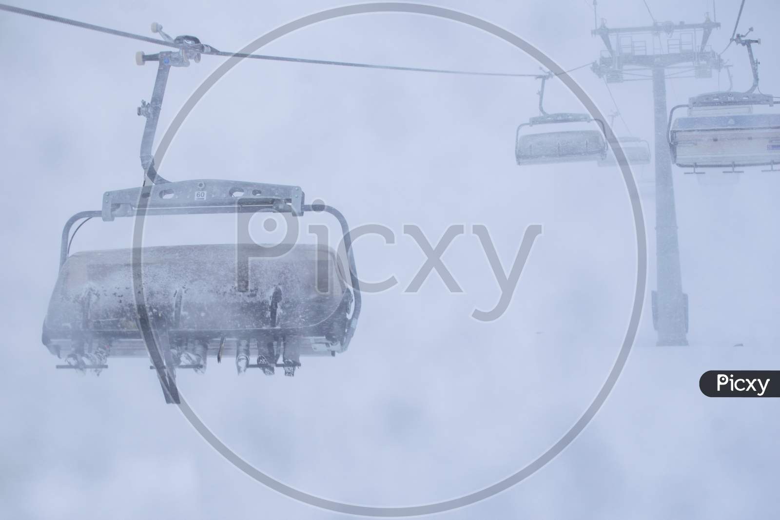 Three Persons In Skis Sit On Ski Resort Chair Lift In Stormy Winter Conditions.Bad Visibility And Weather In Ski Resort Concept.