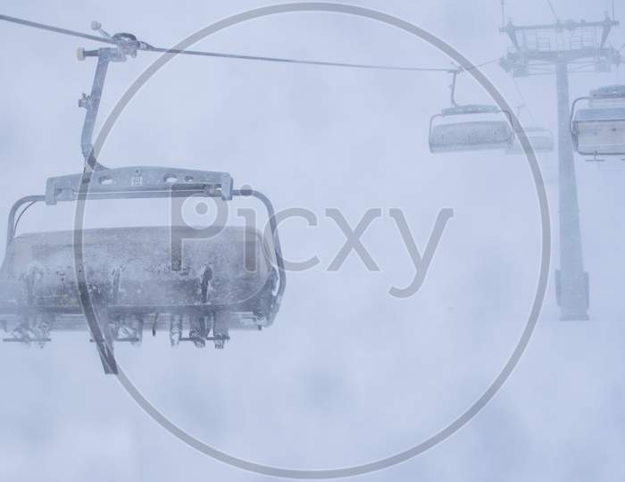 Three Persons In Skis Sit On Ski Resort Chair Lift In Stormy Winter Conditions.Bad Visibility And Weather In Ski Resort Concept.