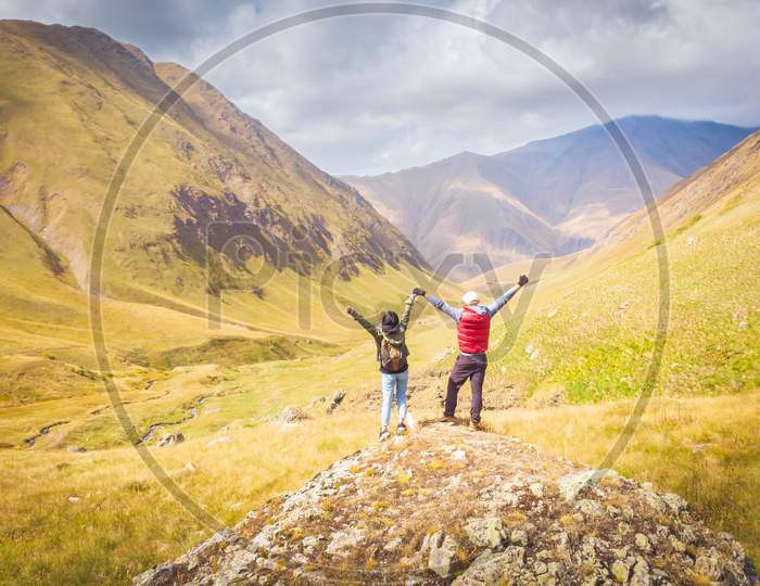 Back Side Of Female And Male On The Rock With Spreaded Hands Up Looking To Beautiful Landscape Of Kazbegi