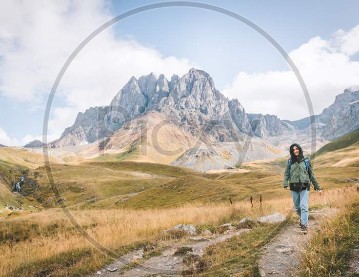 Happy Female Caucasian Woman Walks Down The Path In Juta Valley With Scenic Mountain Landscape. Kazbegi Travel Destination