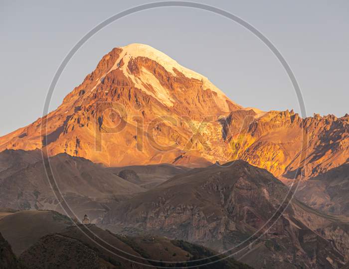 Beaitiful Scenic View Of Kazbek Mountain Top During Sunrise And Gergeti Church In Foreground