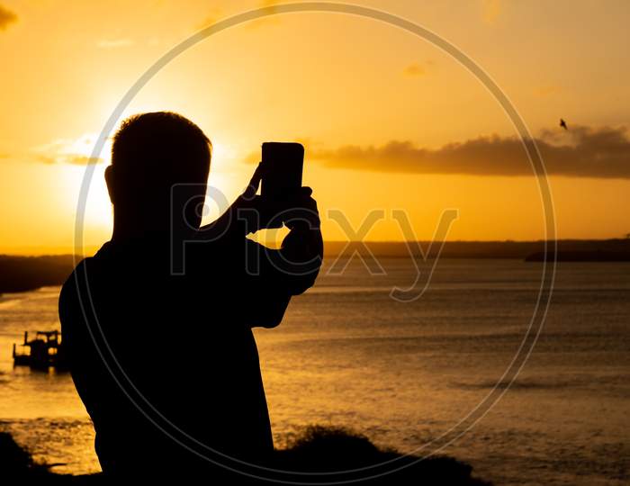 Beautiful And Impressive Sunset On The Edge Of The Lake. Middle-Aged Man Taking A Picture At Sunset With His Smart Phone.