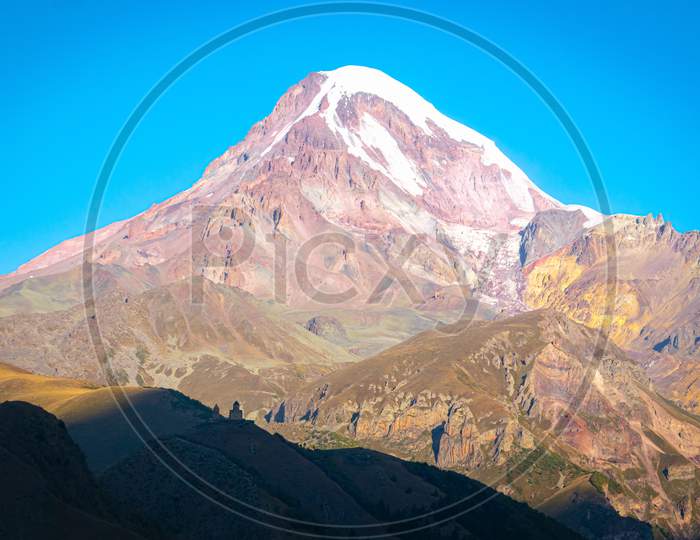 Red Kazbek Mountain Peak With Yellow Hills And Gergeti Trinity Monastery On Shadow In Foreground. Travel Destination In Caucasus.