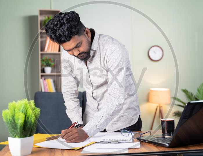Young Business Man Busy In Paperwork By Referring Documents And Laptop - Concept Of Preparing Plan, Managing Tasks By Standing At Office Table.