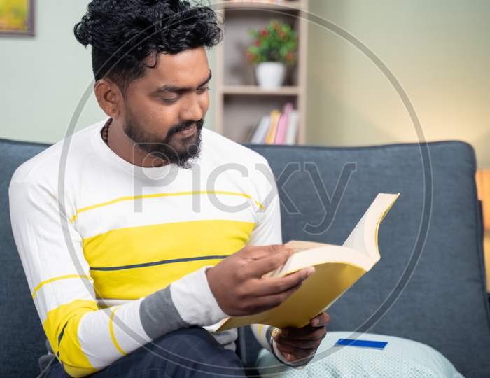 Concentrated Young Man Reading Book During Leisure Time At Home By Sitting On Sofa - Concept Of Reading Hobby.