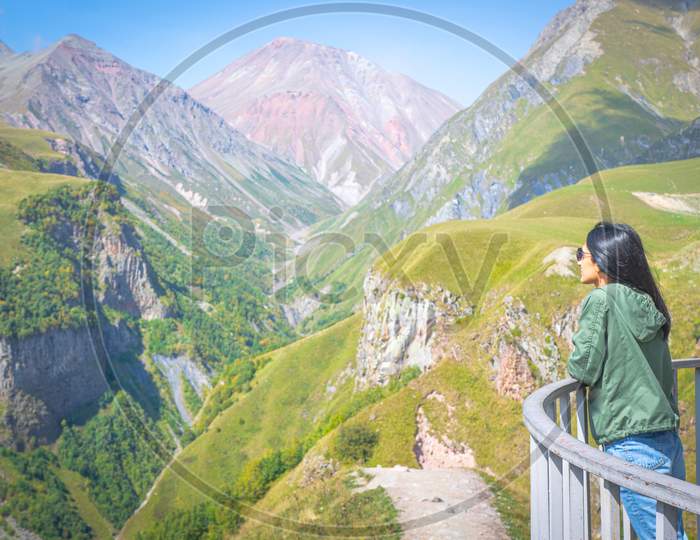 Female Woman Stands On Gudauri Viewpoint Platform And Looks To Scenic Mountain Landscape Of Kazbegi. Travel Destination In Georgia