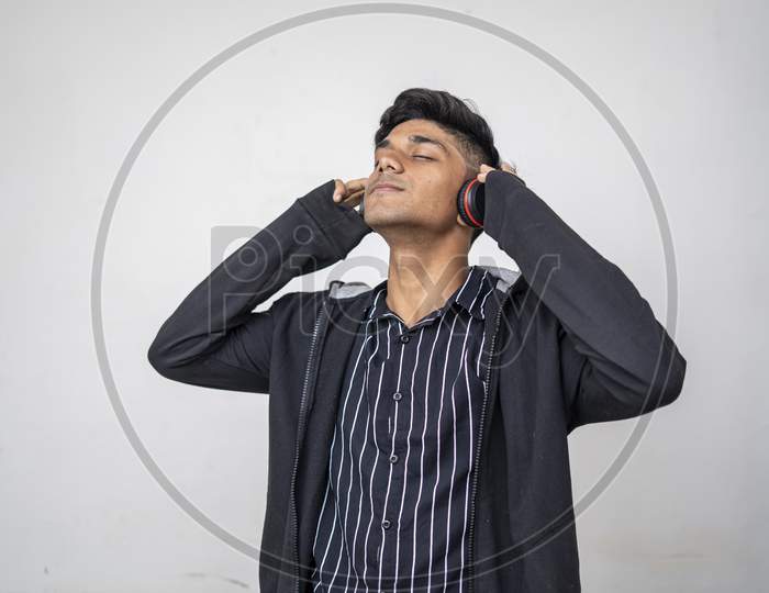 Handsome Attractive Indian Man In Black Hoodie Holding Wireless Music Headphone,Closed Eyes Feel The Music Standing Against White Background.