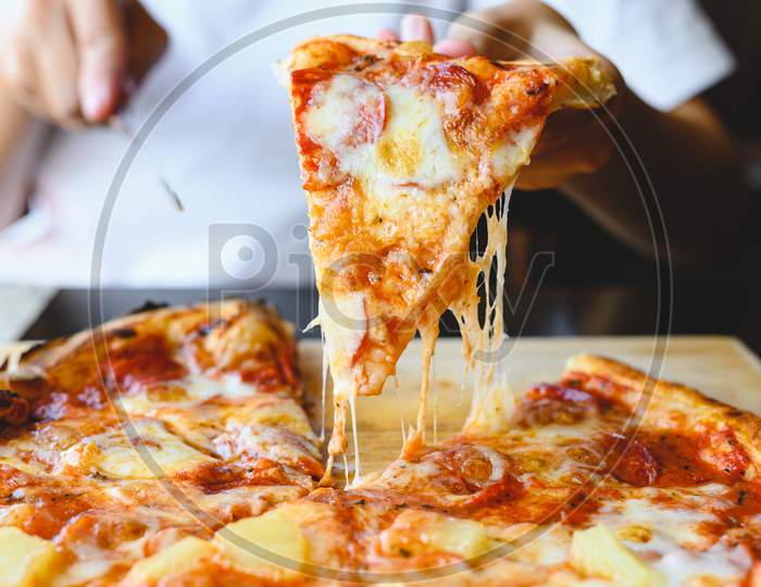 Wood Fire Cheesy Pizza Slice In Hand. Traditional Hawaiian And Salami Pizza Savory Dish Flavour Of Origin Italian Restaurant. Wood-Fired Oven Pizza With People Holding And Cutting Knife Background