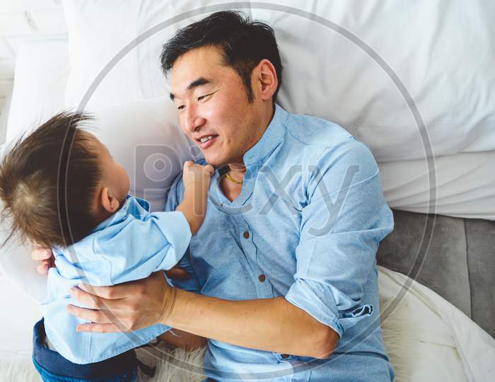 Asian Father And Son Playing Together On White Bed In Bedroom In The Morning. Two People Having Leisure Time At Home. People Lifestyle Concept.