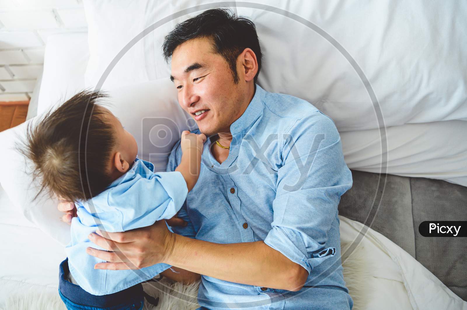 Asian Father And Son Playing Together On White Bed In Bedroom In The Morning. Two People Having Leisure Time At Home. People Lifestyle Concept.