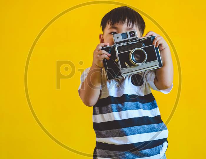 Asian Boy Holding Old Vintage Camera And Posing As Photography On Yellow Isolated Background. People Lifestyle And Technology Concept.