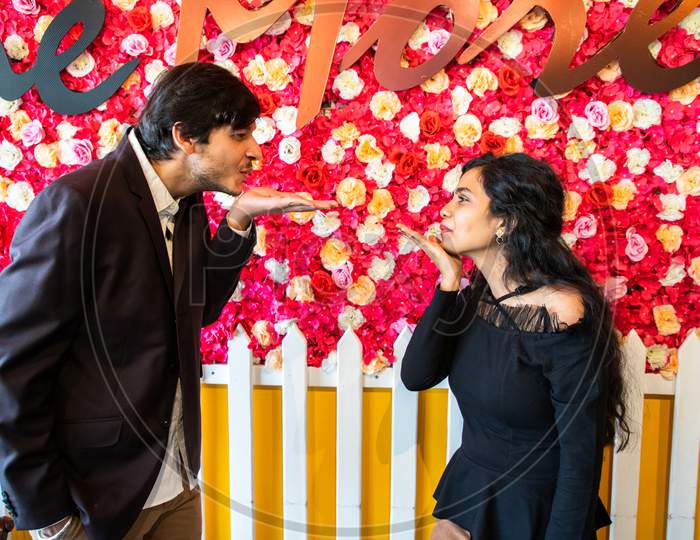 Young Indian Couple In Love Looks At Each Other Blowing Kiss With Hand Or Flying Kiss Against Flower Background. New Year Or Valentines Day Concept.