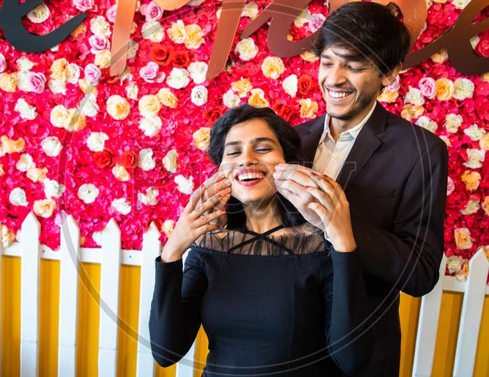 Young Happy Cheerful Indian Couple In Love Have Fun Together, Man Pinching Cheeks Of His Girlfriend Against Floral Background, Enjoy Life, New Year Or Valentines Day.