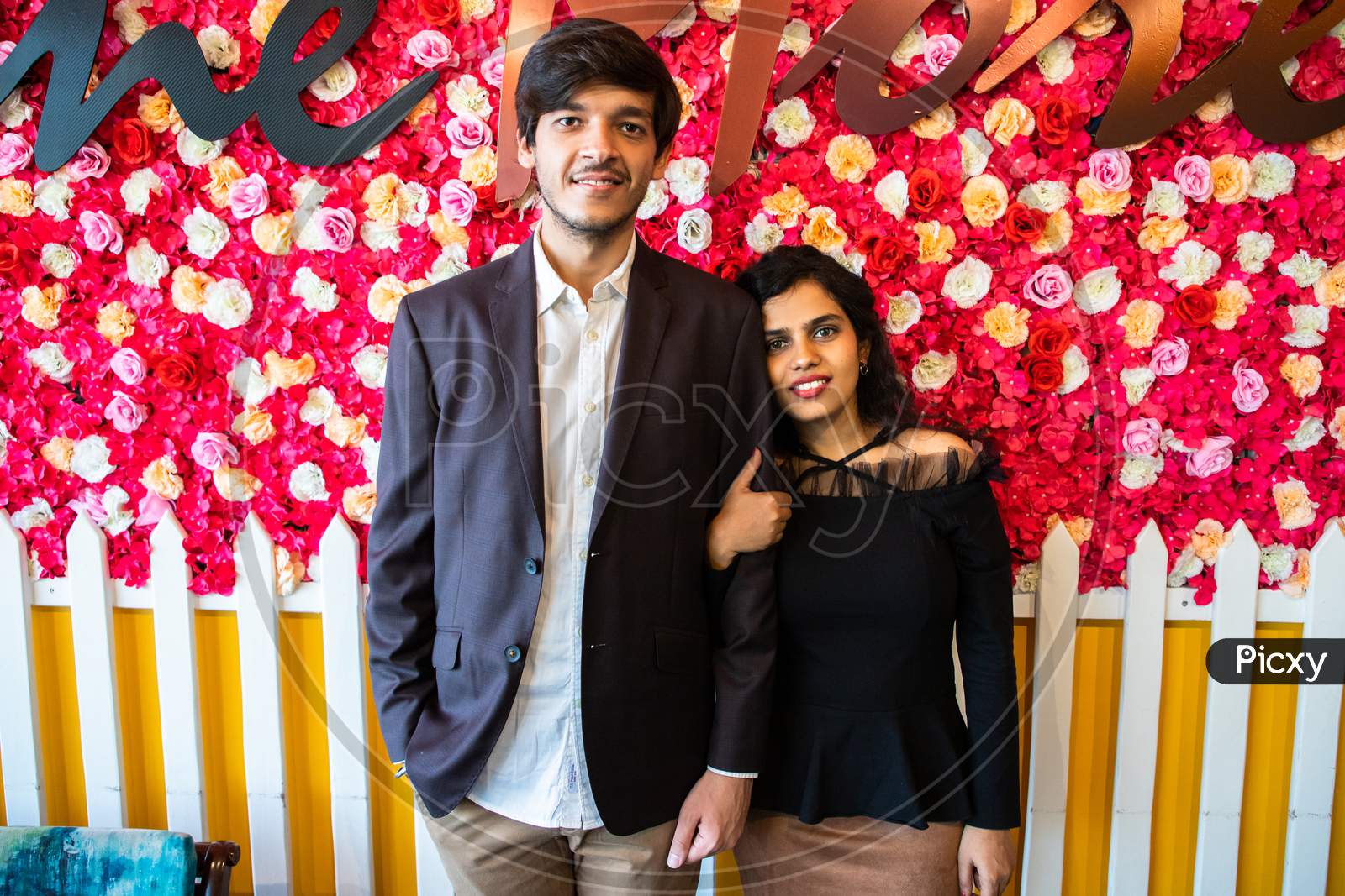 Portrait Of Young Happy Indian Couple In Love Standing Against Floral Background, Boyfriend Girlfriend Relationship, New Year Or Valentines Day.