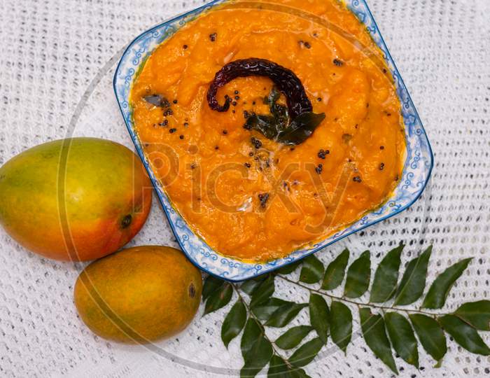 Image of a Goan/konkan mango curry dish made of ripe mangoes on a plate surrounded by mango fruits-perfect for cafe menu