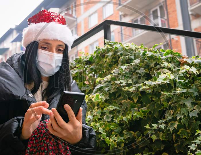 Caucasian Woman With Xmas Hat Texts Outside In The Street. Social Media During Pandemic And Holiday Celebrations And Socail Distancing.