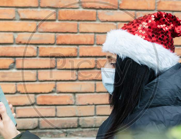 Young Woman Holds Phone Up And Reads Post On Social Media With Xmas Hat On And Brick Wall Blank Space Backgroud. News Media And Social Distancing Duir Corona Virus Pandemic.