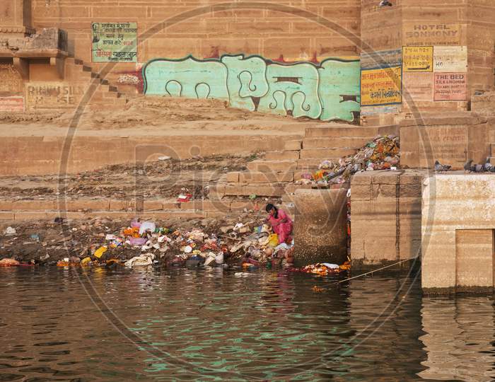 Poor Woman Sits On The Banks Of The Ganges River. Looking For Something To Eat In The Garbage