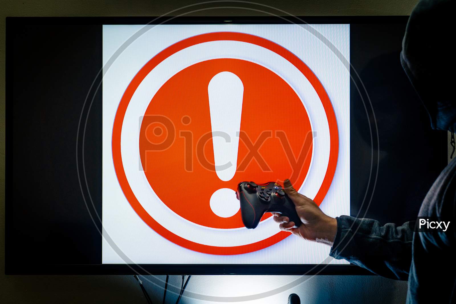 Man In Hood Holding Controller In Front Of Screen Showing An Alert Red Exclamation Message As A Game Crashes Due To Bugs, Hacks Or Issues