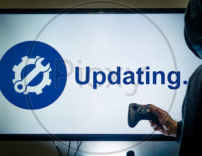 Hooded Man Holding A Gaming Controller In Front Of A Screen Showing An Updating Message For A Game Or Software Day 1 Patch