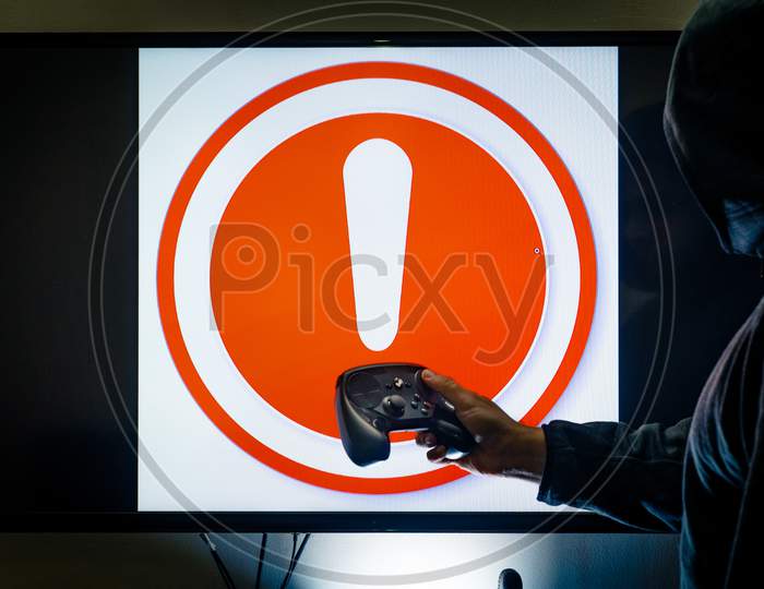 Man In Hood Holding Controller In Front Of Screen Showing An Alert Red Exclamation Message As A Game Crashes Due To Bugs, Hacks Or Issues