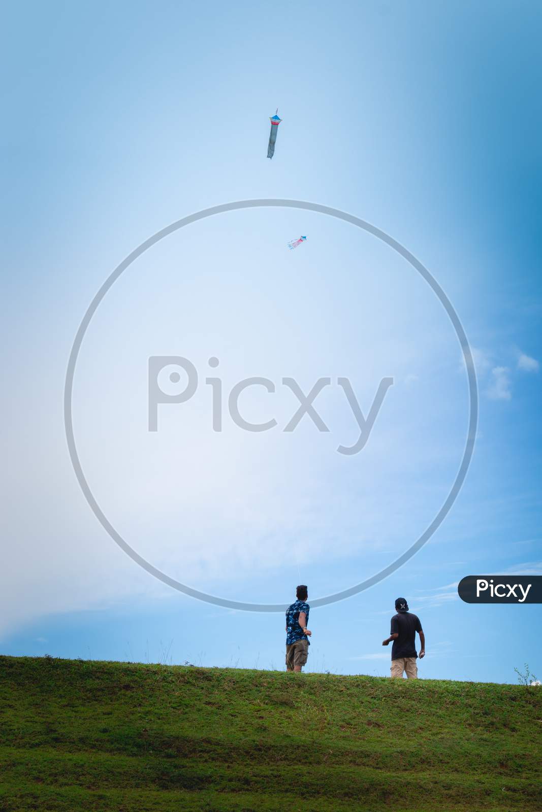 Two Boys Flying Kites In A Grass Land Mountain Hill, Blue Bright Sky
