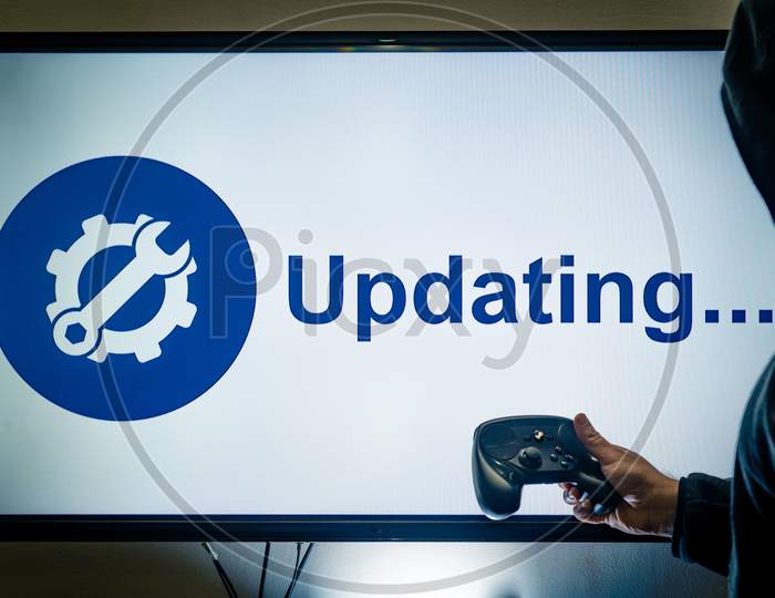Hooded Man Holding A Gaming Controller In Front Of A Screen Showing An Updating Message For A Game Or Software Day 1 Patch