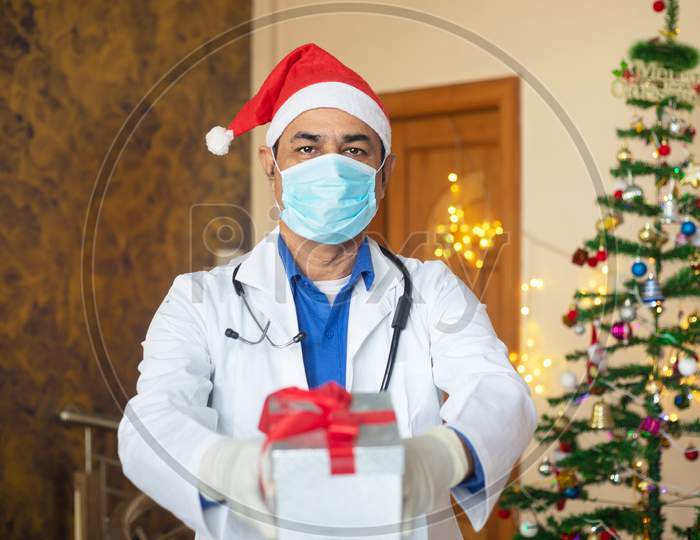 Doctor Wearing Mask And Santa Hat Showing Christmas Gift Box, Celebration During Covid-19 Pandemic, New Normal Lifestyle. Healthcare And Medical.