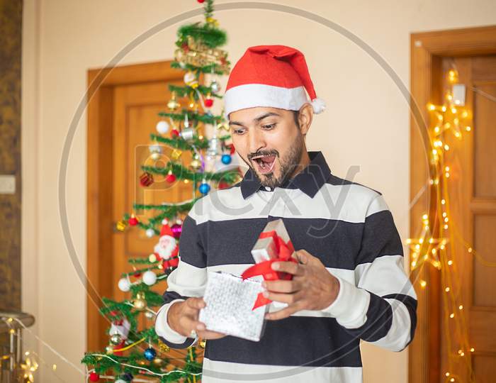 Surprised Amazed Excited Young Man Holding Opened Present Box Wearing Santa Hat At Home, Celebrating Christmas, Holiday.
