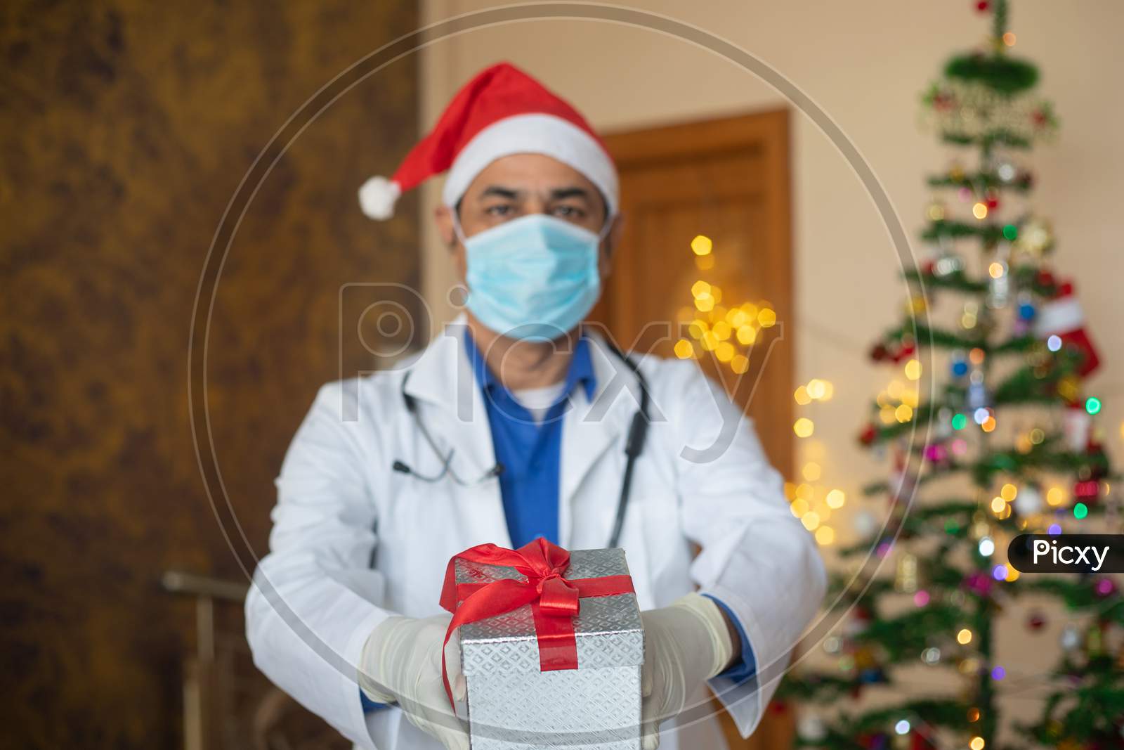 Doctor Wearing Mask And Santa Hat Showing Christmas Gift Box, Celebration During Covid-19 Pandemic, New Normal Lifestyle. Healthcare And Medical, Selective Focus.