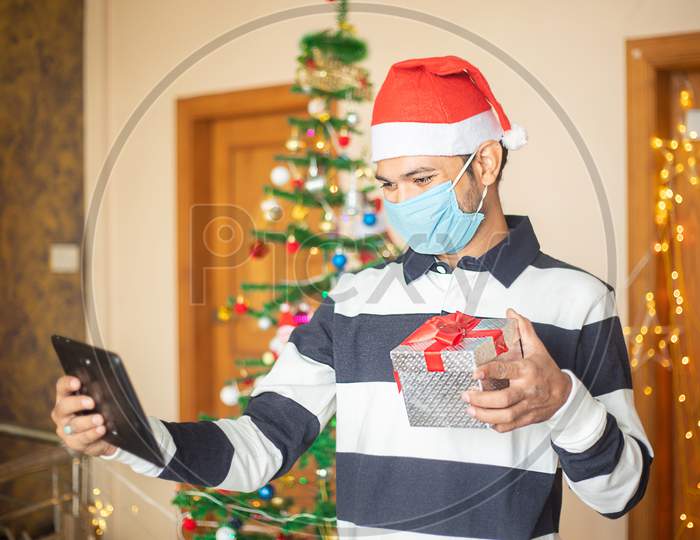 Online Christmas Celebration, Young Man Wearing Mask Congratulating Over Video Calling On Tablet Showing Gift Present, Covid-19 Pandemic, New Year, Holiday,Winter,December,New Normal.