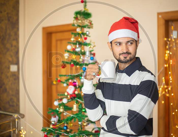Young Man Wearing Santa Hat Holding Coffee Mug In Hand Celebrating Christmas Alone At Home, New Year, Holiday, Party, Winter, December. Quarantine Due To Covid-19 Pandemic.