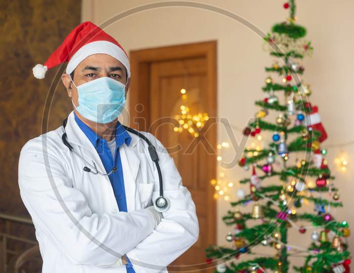 Portrait Of Doctor Wearing Mask And Christmas Hat Celebration During Covid-19 Pandemic, Lockdown, New Normal. Healthcare And Medical Person Looking At Camera With Decorated Tree In The Background