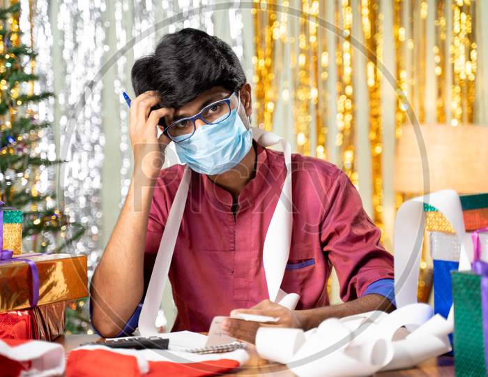 Young Man In Face Mask Worried About Holyday Expenses After Christmas Or New Year 2021 Holiday Celebration During Coronavirus Covid-19 Pandemic With Gift And Bills In Front.