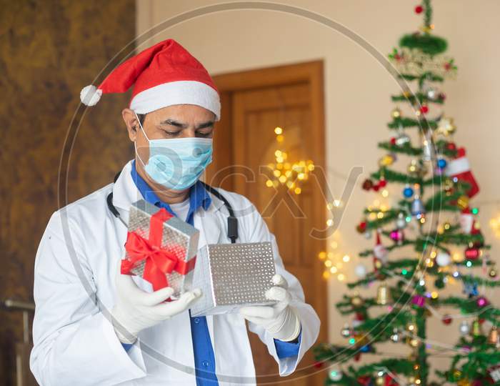 Doctor Wearing Mask And Santa Hat Open Christmas Gift Box, Celebration During Covid-19 Pandemic, New Normal Lifestyle. Healthcare And Medical. Decorated Tree In The Background