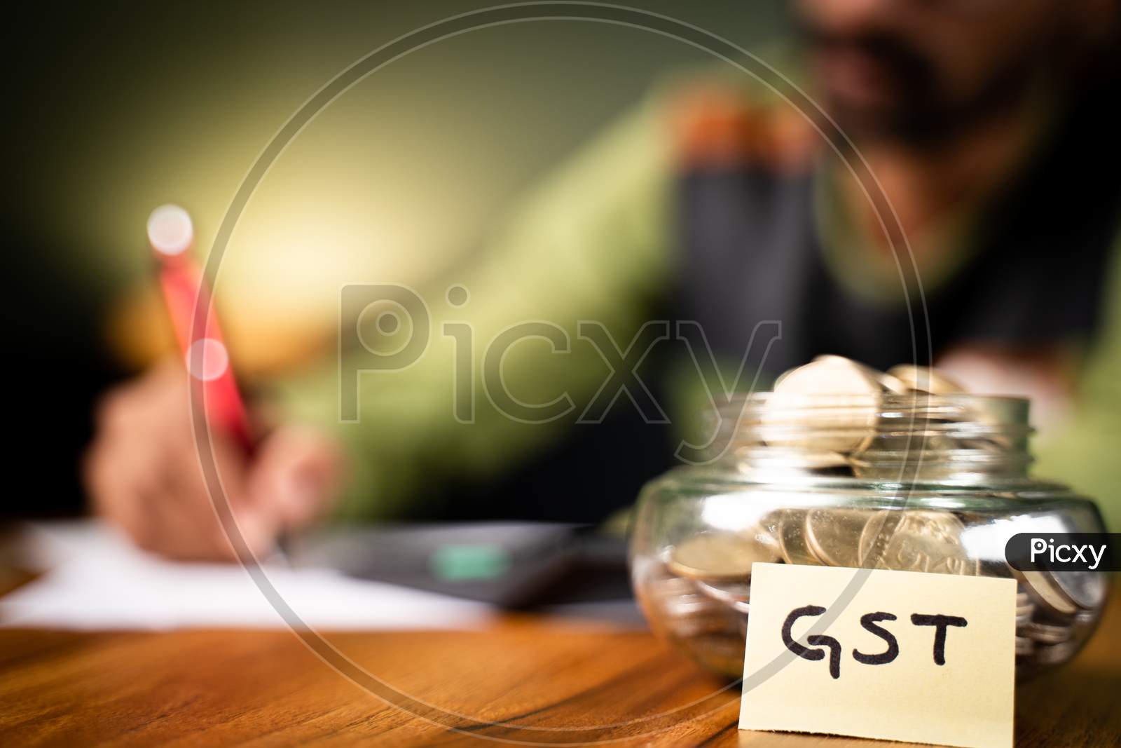 Selective Focus On Coins In Bottle And Gst Sticky Note, Man In Background Checking Spendings On Calculator - Concept Of Gst Tax Savings Calculation.