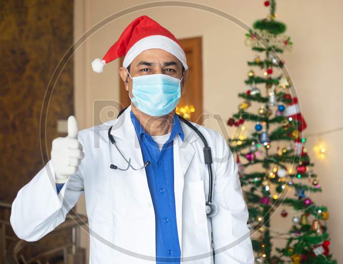 Doctor Wearing Mask And Christmas Santa Hat Doing Thumb Up, Healthcare Medical Person On Duty Celebration During Covid-19 Pandemic, Lockdown, New Normal.