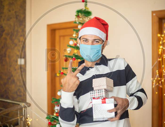 Cheerful Young Man With Santa Hat Holding Lots Of Christmas Gift Boxes, Male Model Wearing Mask Celebrating During Covid-19 Pandemic.