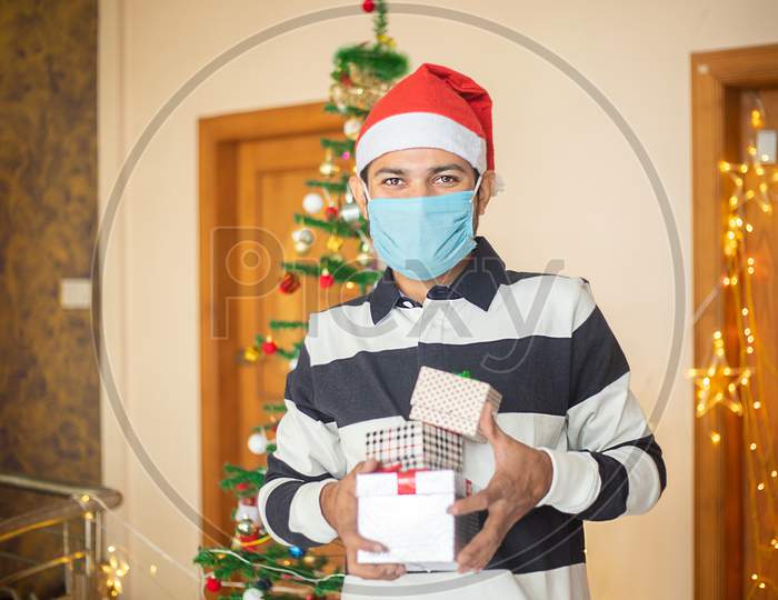 Young Man Wearing Mask And Santa Hat Holding Lots Of Christmas Gift Boxes, Celebrating During Covid-19 Pandemic, New Year, Holiday,Winter,December,New Normal.