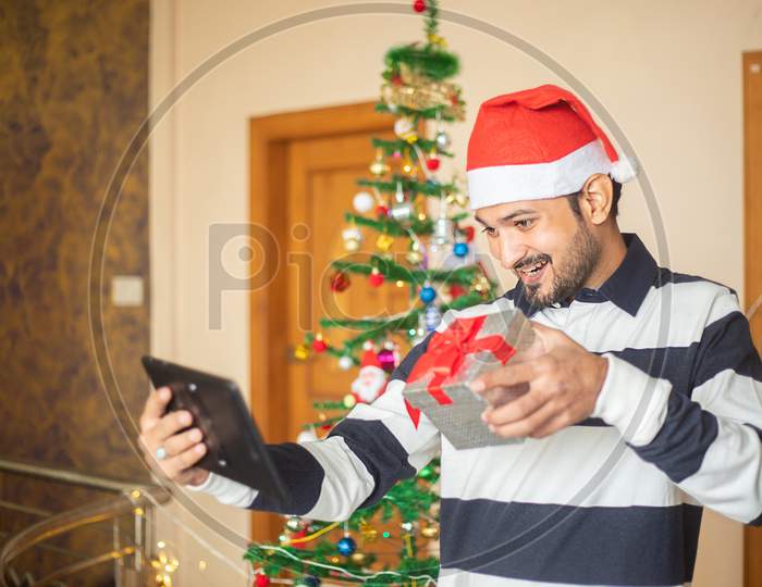 Online Christmas Celebration, Young Man  Congratulating Over Video Calling On Tablet Showing Gift Present, Covid-19 Pandemic, New Year, Holiday,Winter,December,New Normal.