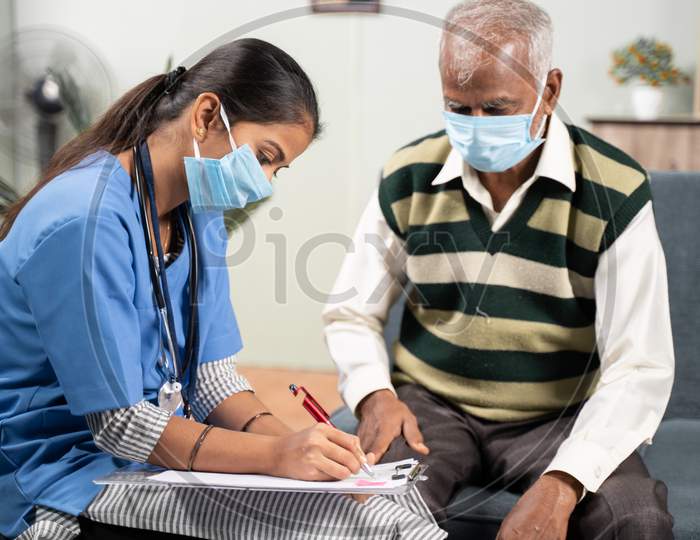 Young Doctor Or Nurse Writing Prescription During Home Visiting To Sick Elder Man While Both Worn Face Mask Due To Coronavirus Covid-19 Pandemic.