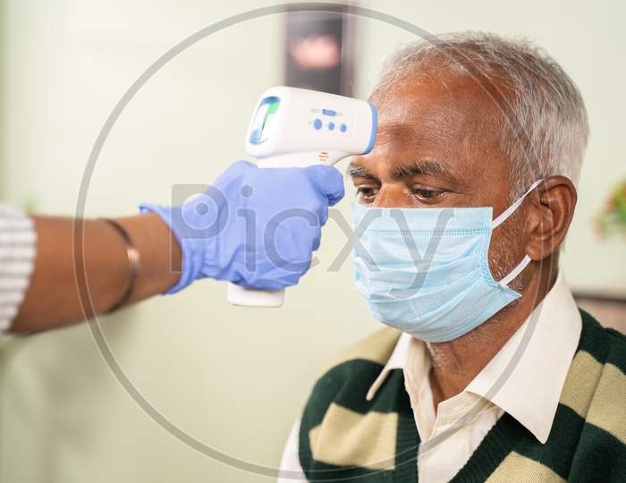 Close Up Of Doctor Hands Checking Temperature Of Old Sick Man Using Infrared Thermometer At Hospital While Both Worn Face Mask Due To Coronavirus Covid-19 Safety Measures.