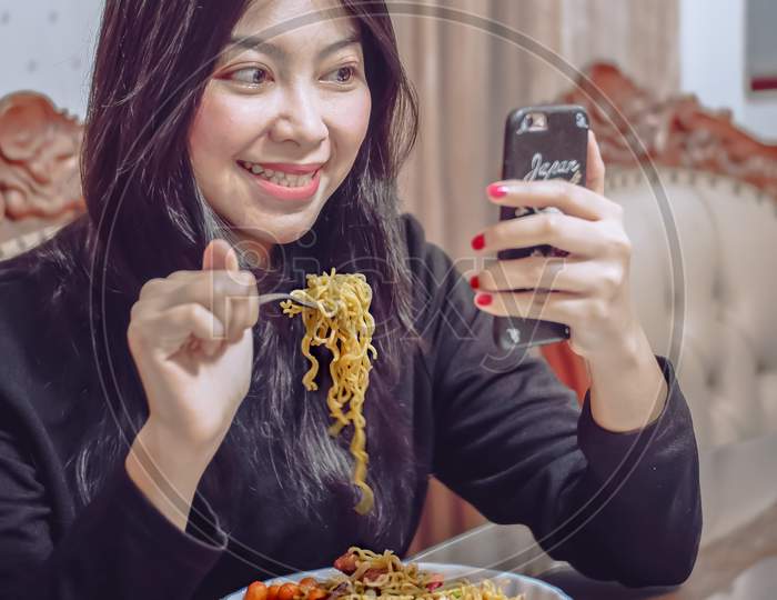 Funny cute asian girl looking to mobile phone in her hand while eating noodles
